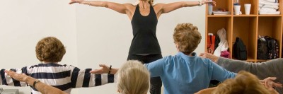 Female instructor leading a “chair yoga” class with female patients seated with back to camera and arms stretched to side.